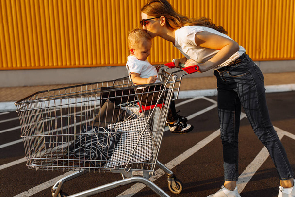 Attractive happy young woman in sunglasses shopping with small child, trolley and shopping bags, outdoors - Zdjęcie, obraz