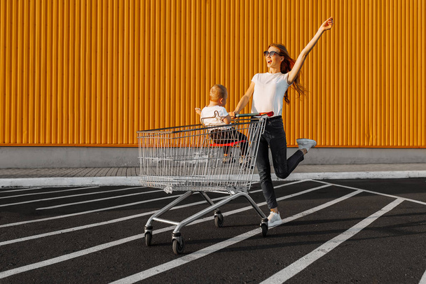 Happy excited young woman with baby and trolley on yellow wall of shopping mall, family shopping - 写真・画像