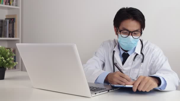 Young Asian Doctor Man in Lab Coat or Gown with Stethoscope Wear Glasses and Face Mask Exam Patient by Video Call or Video Conference in Office - Footage, Video