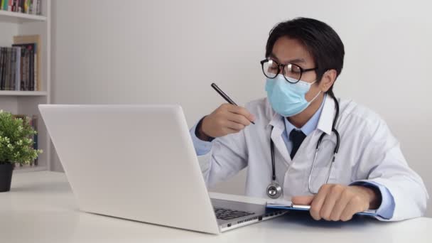 Young Asian Doctor Man in Lab Coat or Gown with Stethoscope Wear Glasses and Face Mask Suggest and Exam Patient by Video Call or Video Conference in Office - Footage, Video