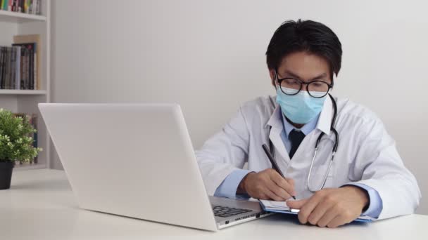 Young Asian Doctor Man in Lab Coat or Gown with Stethoscope Wear Glasses and Face Mask Writing Patient Exam Report in Office - Footage, Video