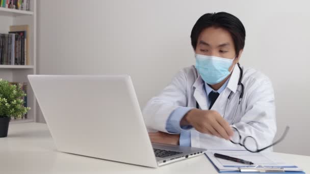 Young Asian Doctor Man in Lab Coat or Gown with Stethoscope Wear Face Mask and Holding Glasses Using Laptop in Office - Footage, Video