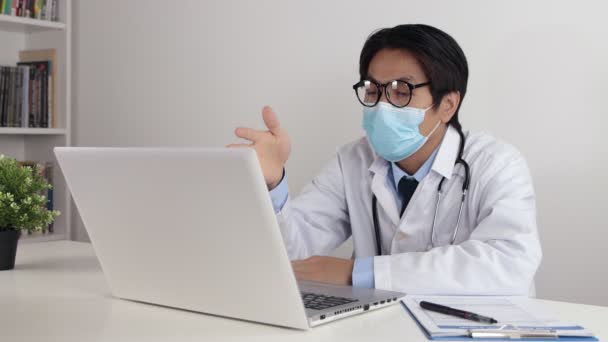 Young Asian Doctor Man in Lab Coat or Gown with Stethoscope Wear Face Mask and Glasses Exam Patient by Video Conference or Video Chat in Office - Footage, Video
