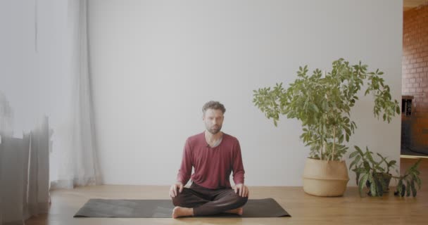 Kaukasischer Mann sitzt im Schneidersitz und meditiert mit geschlossenen Augen drinnen in einer sonnigen Wohnung. Porträt eines bärtigen Yoga-Trainers, der sich in Sukhasana Asana Zeitlupe entspannt. Entspannungsstress überwunden - Filmmaterial, Video