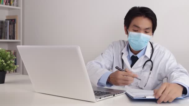 Young Asian Doctor Man in Lab Coat or Gown with Stethoscope Wear Face Mask Exam Patient by Video Chat or Video Conference in Office - Footage, Video