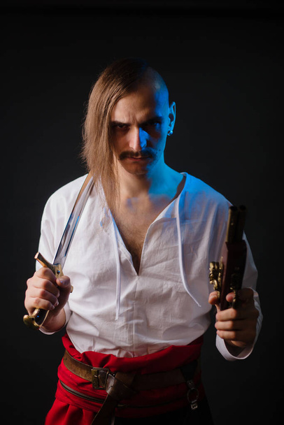 Kozak with weapons in national Ukrainian clothes. A young man with a forelock, in a white shirt and trousers with a red belt, holds a saber and a pistol. - Photo, Image