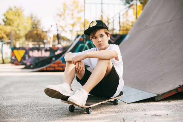 Красивый подросток сидит на скейтборде в специальной зоне парка. Мальчик отдыхает после поездки в скейтпарке. Активный отдых на свежем воздухе - Фото, изображение