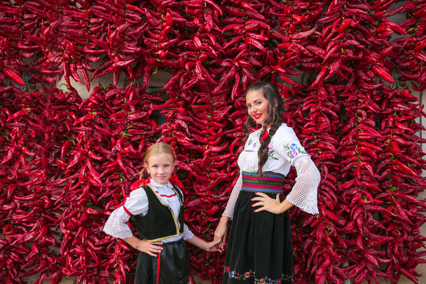 Κορίτσια ντυμένα με παραδοσιακά σερβικά βαλκανικά ρούχα, εθνική λαϊκή ενδυμασία. Τοποθέτηση κοντά σε παρτίδα κόκκινες πιπεριές πάπρικα κρέμεται στον τοίχο του σπιτιού. . - Φωτογραφία, εικόνα