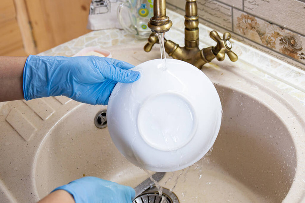 Ręce kobiet w niebieskich rękawiczkach zmywają naczynia i talerze w zlewie kuchennym. Koncepcja gospodarstwa domowego - Zdjęcie, obraz
