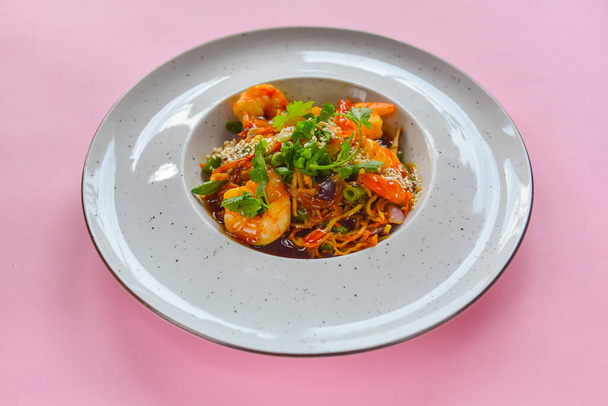 Asian noodles with shrimps, asparagus and sesame seeds. Delicious shrimp wog, traditional Asian cuisine. Served in a white plate over pastel pink background. Copy space banner. - Foto, afbeelding
