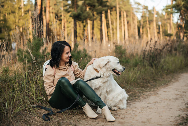 brunette girl sitting with white golden retriever dog on the forest path - Foto, afbeelding