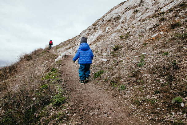 Dziecko w niebieskiej kurtce idzie ścieżką. Mały chłopiec wspina się na górę sam. Trekking na świeżym powietrzu dla dzieci w przyrodzie - Zdjęcie, obraz