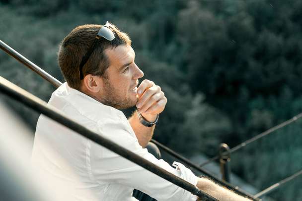 Portré egy fiatal, vonzó férfiról fehér ingben, aki ül és a gyönyörű zöld hegyek hátterét nézi. - Fotó, kép