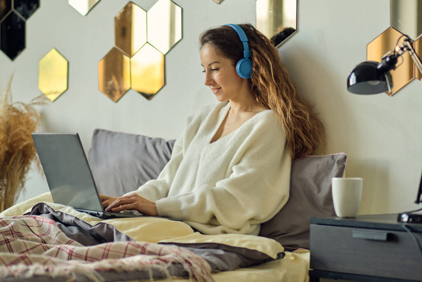Νεαρή όμορφη γυναίκα που φοράει ακουστικά ενώ χρησιμοποιεί φορητό υπολογιστή στο κρεβάτι - Φωτογραφία, εικόνα