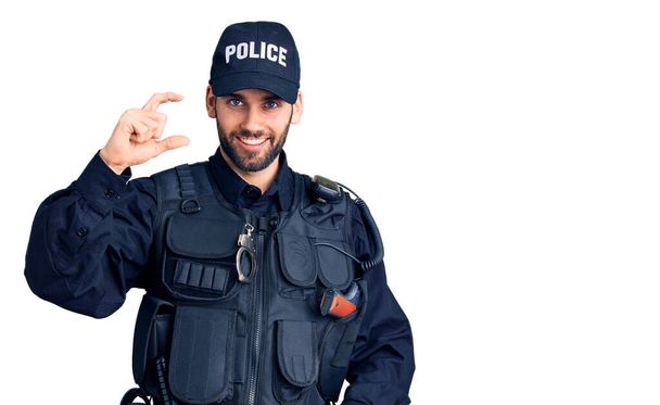 Молодий красивий чоловік з бородою в поліцейській формі посміхається і впевнено жестикулює рукою, роблячи знак малого розміру з пальцями, дивлячись і камеру. міра концепції
.  - Фото, зображення