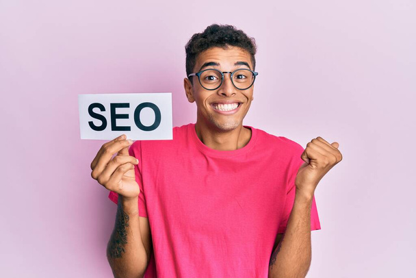 Young handsome african american man holding seo message paper screaming proud, celebrating victory and success very excited with raised arm  - Photo, Image