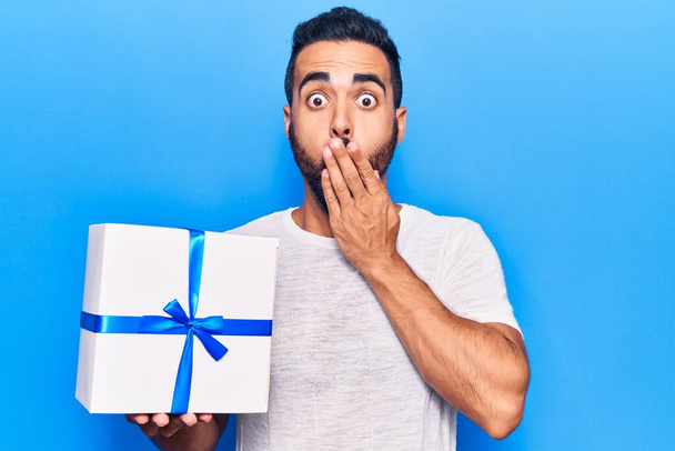 Young hispanic man holding gift covering mouth with hand, shocked and afraid for mistake. surprised expression  - Foto, Bild