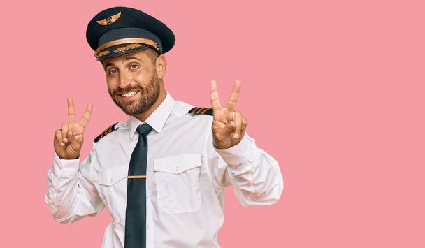 Hombre guapo con barba llevando uniforme de piloto de avión sonriendo mirando a la cámara mostrando los dedos haciendo señal de victoria. número dos.  - Foto, imagen