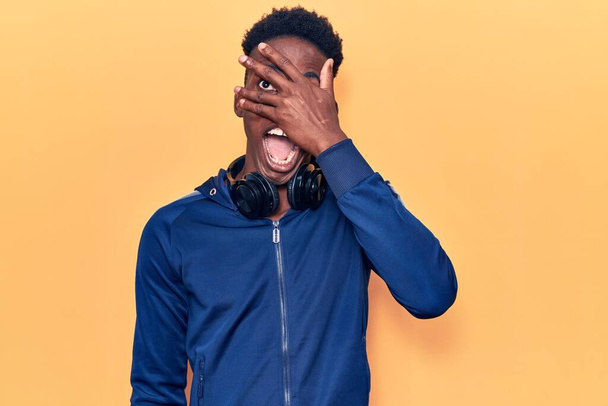 Jeune homme afro-américain portant des vêtements de sport et des écouteurs regardant dans le choc couvrant le visage et les yeux avec la main, regardant à travers les doigts peur  - Photo, image