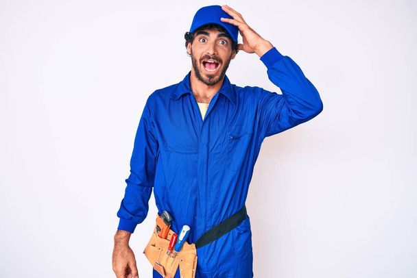 Handsome young man with curly hair and bear weaing handyman uniform surprised with hand on head for mistake, remember error. forgot, bad memory concept.  - Photo, Image
