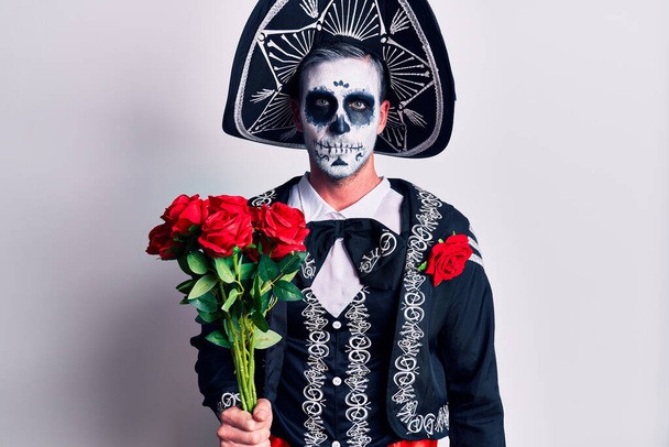Νεαρός άνδρας φορώντας μεξικάνικη ημέρα του νεκρού κοστούμι κρατώντας λουλούδια σκέψης στάση και νηφάλια έκφραση που αναζητούν αυτοπεποίθηση  - Φωτογραφία, εικόνα
