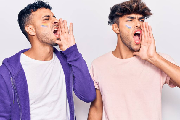 Joven pareja gay dibujando arco iris lgtbq bandera en la cara gritando y gritando fuerte a un lado con la mano en la boca. concepto de comunicación.  - Foto, Imagen