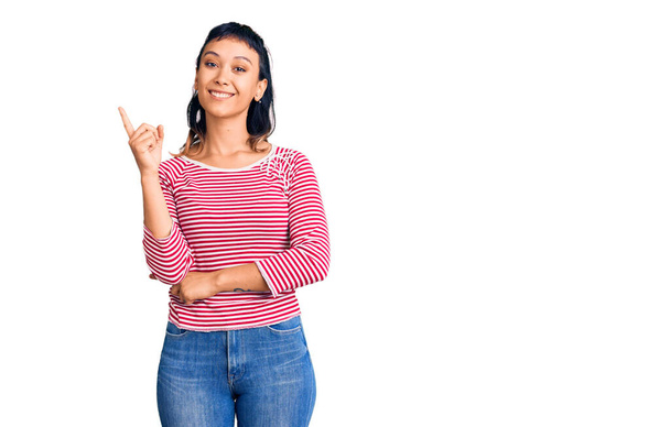 Młoda kobieta w luźnych ubraniach uśmiecha się szczęśliwa wskazując ręką i palcem na bok  - Zdjęcie, obraz