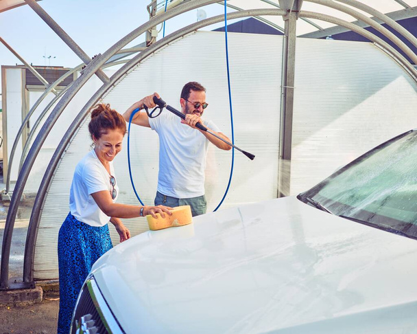 Middle age beautiful couple wearing casual clothes and smiling happy. Standing with smile on face washing car using water pistol and sponge. - Photo, Image