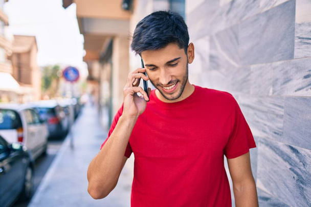 Młody Latynos uśmiecha się szczęśliwy rozmawiając na smartfonie spacerując po mieście. - Zdjęcie, obraz