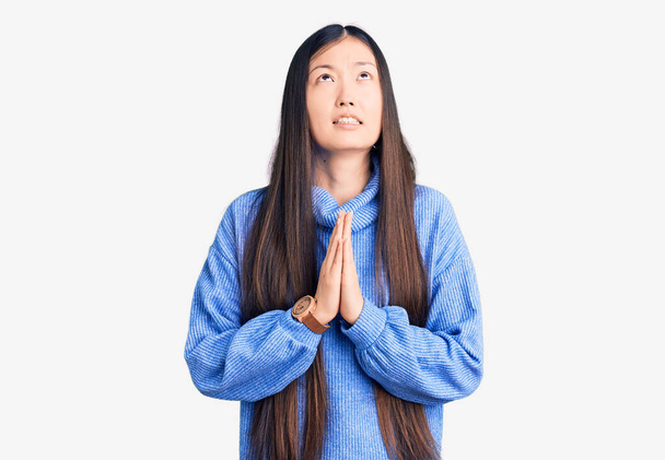 Jovem mulher chinesa bonita vestindo camisola de gola alta casual implorando e orando com as mãos, juntamente com a expressão de esperança no rosto muito emocional e preocupado. implorando.  - Foto, Imagem