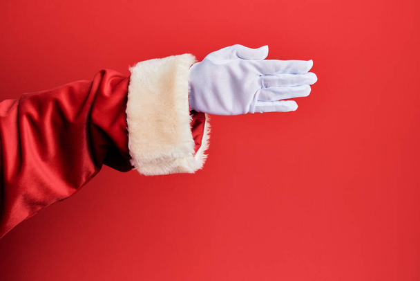 Main d'un homme portant costume santa claus et gants sur fond rouge étirant et atteignant avec la main ouverte pour poignée de main, montrant la paume  - Photo, image