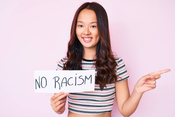Νεαρή όμορφη Κινέζα κοπέλα που δεν κρατά πανό ρατσισμού χαμογελώντας χαρούμενη δείχνοντας με το χέρι και το δάχτυλο στο πλάι  - Φωτογραφία, εικόνα
