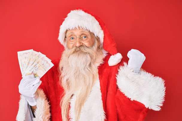 Старик с седыми волосами и длинной бородой в костюме Санта-Клауса держит фунты, гордо крича, празднуя победу и успех очень взволнованный с поднятой рукой  - Фото, изображение
