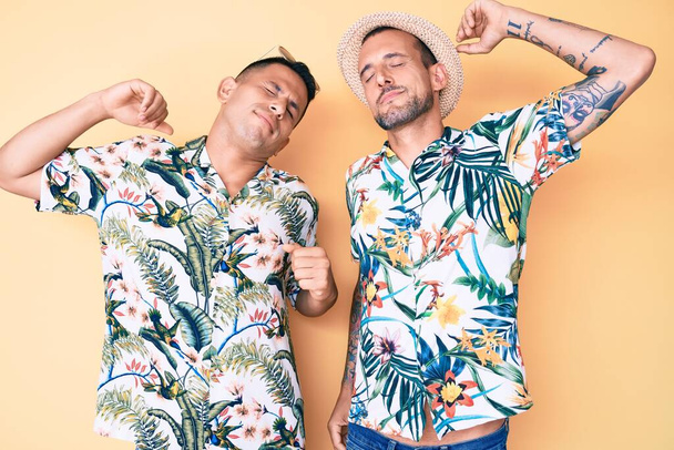 młody gej para z dwa mężczyźni nosić letni kapelusz i hawajski koszula rozciągający się z powrotem zmęczony i zrelaksowany senny i ziewanie dla wczesny ranek  - Zdjęcie, obraz