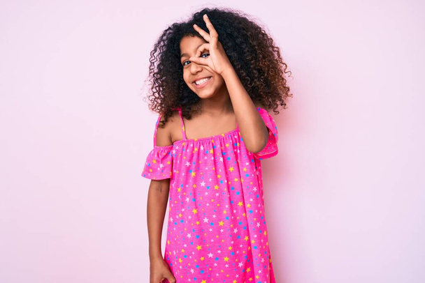 African american child with curly hair wearing casual dress doing ok gesture with hand smiling, eye looking through fingers with happy face.  - Foto, immagini
