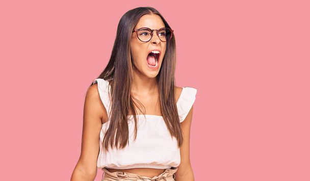 Young hispanic woman wearing casual clothes and glasses angry and mad screaming frustrated and furious, shouting with anger. rage and aggressive concept.  - Photo, image