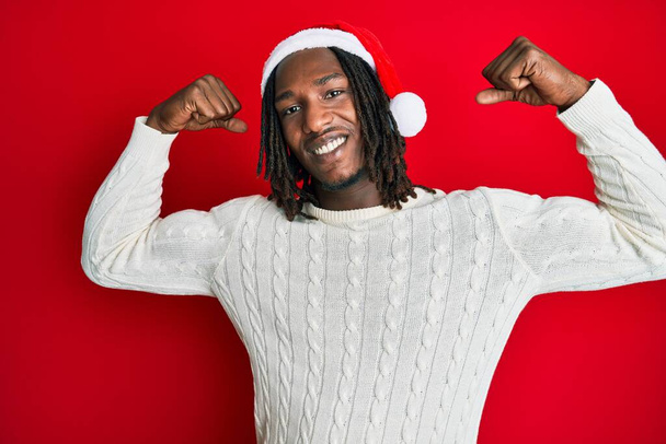 Αφροαμερικάνος με κοτσίδες που φοράει χριστουγεννιάτικο καπέλο και δείχνει μπράτσα να χαμογελούν περήφανοι. έννοια της καταλληλότητας.  - Φωτογραφία, εικόνα
