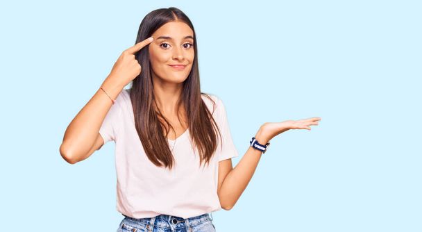 Jonge Latijns-Amerikaanse vrouw draagt casual witte tshirt verward en geïrriteerd met open palm tonen kopieerruimte en wijzende vinger naar het voorhoofd. Denk er eens over na..  - Foto, afbeelding