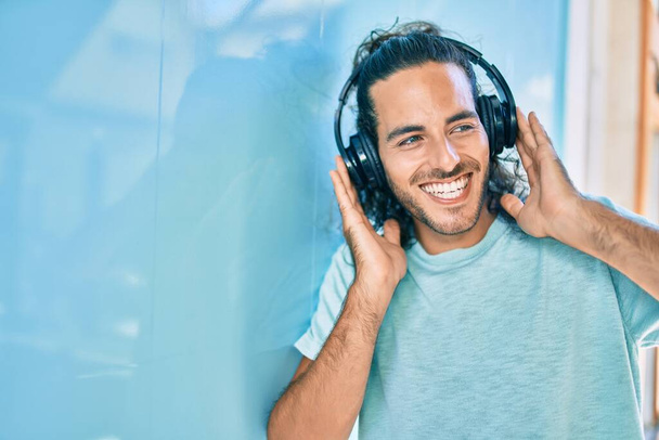 Młody Latynos uśmiechający się szczęśliwy słuchając muzyki za pomocą słuchawek w mieście. - Zdjęcie, obraz