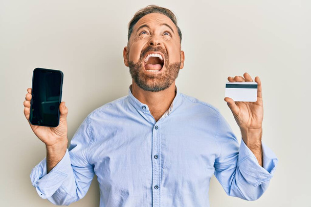 Guapo hombre de mediana edad sosteniendo teléfono inteligente y tarjeta de crédito enojado y loco gritando frustrado y furioso, gritando con ira mirando hacia arriba.  - Foto, imagen