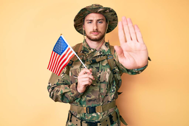 Joven hombre caucásico con camuflaje uniforme del ejército sosteniendo bandera de EE.UU. con la mano abierta haciendo señal de stop con expresión seria y segura, gesto de defensa  - Foto, Imagen