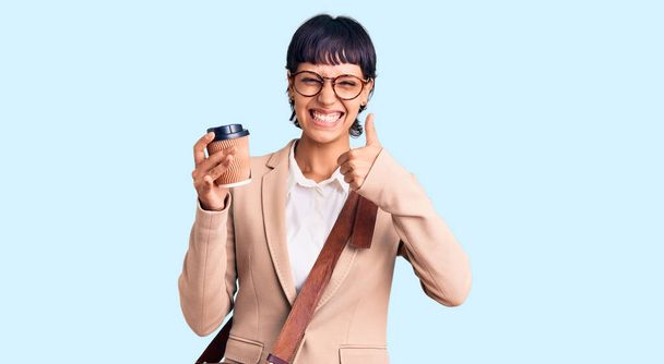 Молодая брюнетка женщина с короткими волосами носить деловую сумку и пить кофе улыбаясь счастливый и позитивный, большой палец вверх делает отлично и знак одобрения  - Фото, изображение