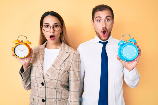Beautiful couple wearing business jacket and holding alarm clock afraid and shocked with surprise and amazed expression, fear and excited face.  - Photo, Image