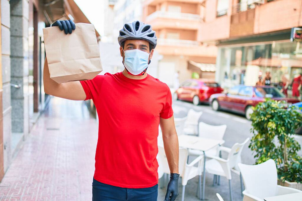 若い配達人は自転車のヘルメットとコロナウイルス保護医療用マスクを着て街の通りで紙袋を奪う. - 写真・画像