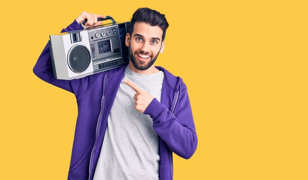 Young handsome man with beard listening to music using vintage boombox smiling happy pointing with hand and finger  - Photo, Image