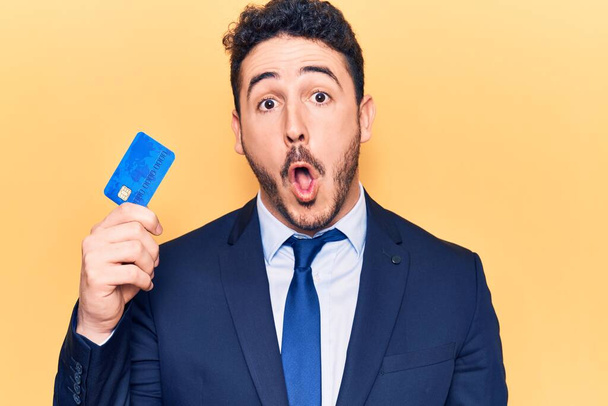 Jovem hispânico vestindo terno segurando cartão de crédito assustado e surpreso com a boca aberta para surpresa, rosto descrença  - Foto, Imagem