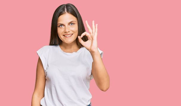 Молодая кавказская женщина в обычной белой футболке, улыбаясь позитивно, делает хорошо знак с рукой и пальцами. успешное выражение.  - Фото, изображение