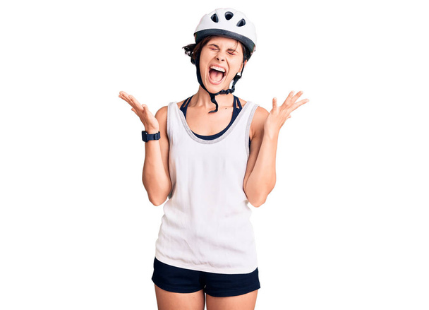 Beautiful young woman with short hair wearing bike helmet celebrating mad and crazy for success with arms raised and closed eyes screaming excited. winner concept  - Photo, Image
