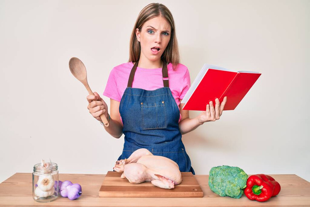 Νεαρό ξανθό κορίτσι φορώντας επαγγελματική ποδιά φούρναρη ανάγνωση βιβλίο συνταγών μαγειρικής στο πρόσωπο σοκ, αναζητούν δύσπιστοι και σαρκαστικός, έκπληκτος με ανοιχτό στόμα  - Φωτογραφία, εικόνα