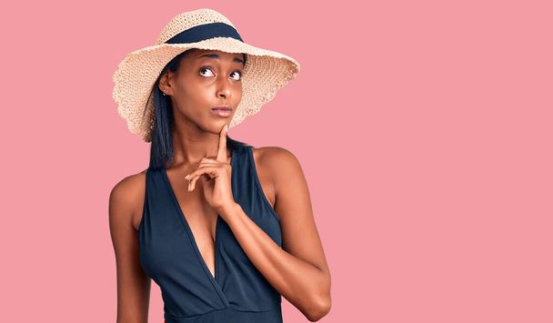 Νεαρή Αφροαμερικανή γυναίκα που φοράει μαγιό και καλοκαιρινό καπέλο σκέφτεται συγκεντρωμένα την αμφιβολία με το δάχτυλο στο πηγούνι και αναρωτιέται  - Φωτογραφία, εικόνα